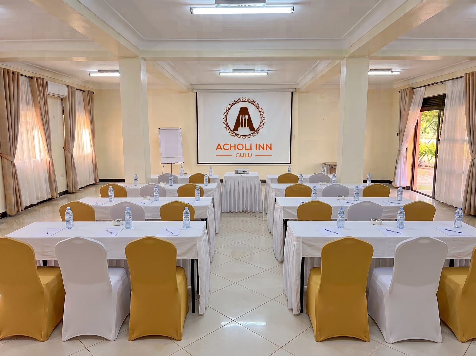 Karuma Hall set up classroom-style with rows of skirted tables, gold and white chairs and the Acholi Inn logo on the projector screen